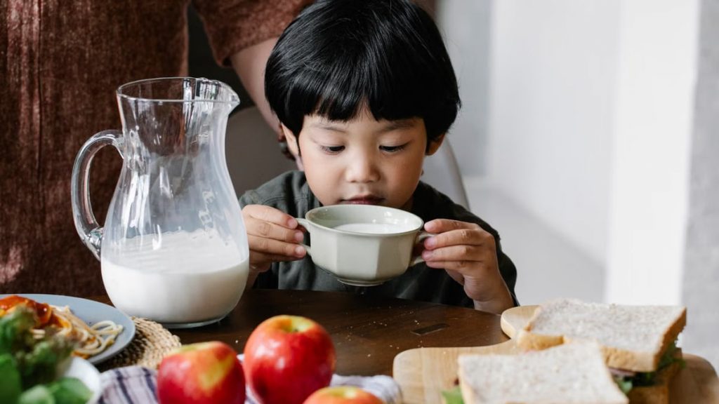 Asian little boy drinking milk from mug while sitting at served table in kitchen with crop anonymous woman behind back